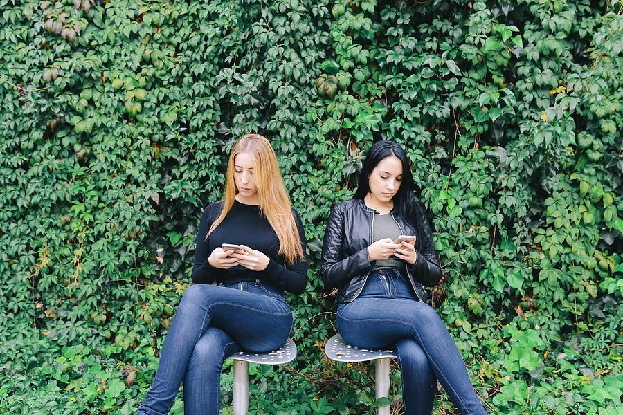 Two young women sitting side by side using their cell phones Photograph by Westend61
