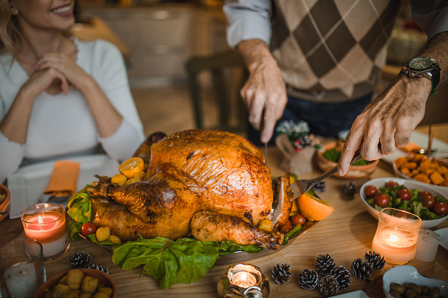 Unrecognizable person carving roasted turkey during Thanksgiving dinner at dining table. Photograph by Skynesher