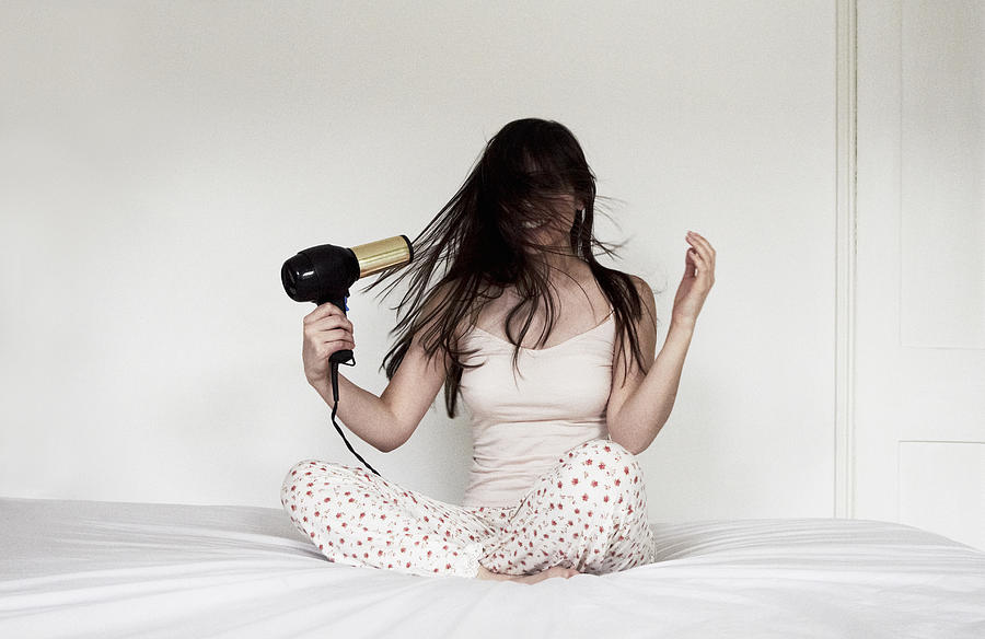Woman sitting on bed blow drying hair Photograph by Flashpop