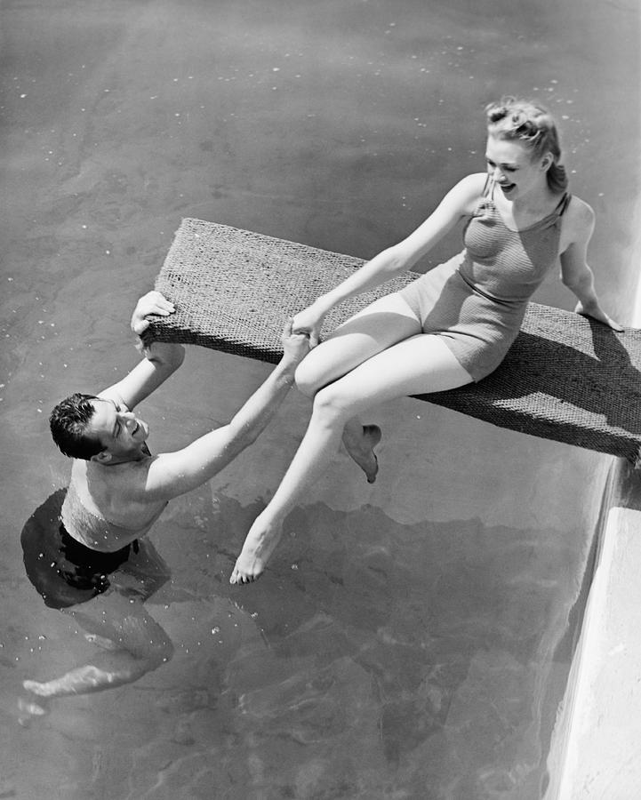 Woman sitting on diving board, man grasping her hand (B&W), elevated view Photograph by George Marks