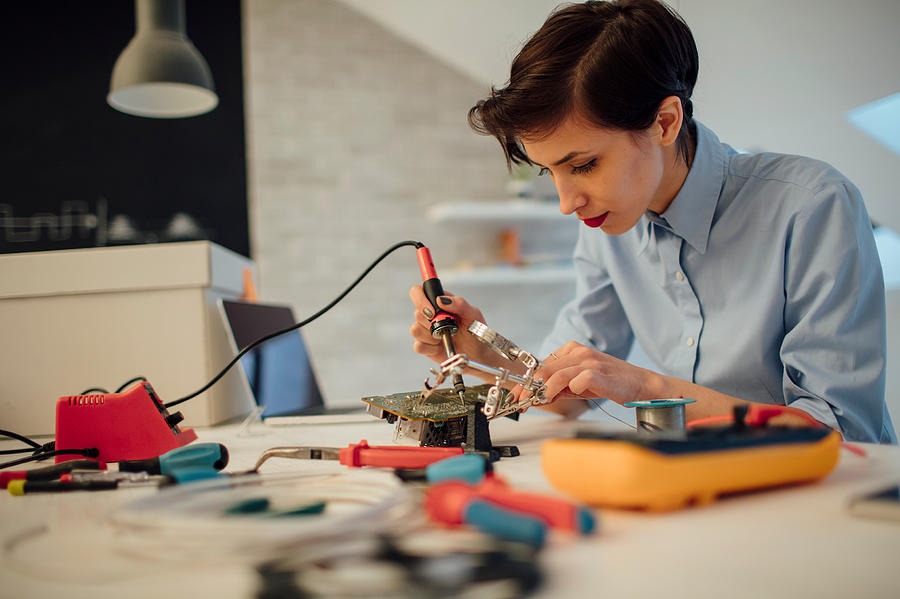 Woman Soldering a circuit board in her tech office. Photograph by Vgajic