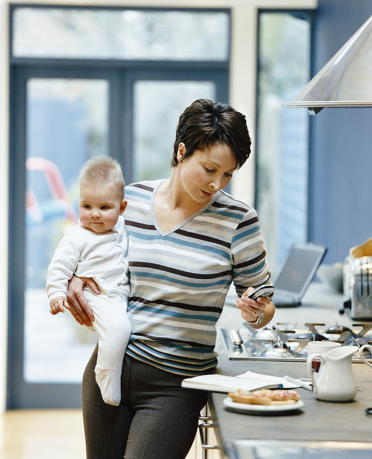 Woman Stands by a Kitchen Counter Holding Her Baby and Dialing Her Mobile Phone Photograph by Digital Vision.
