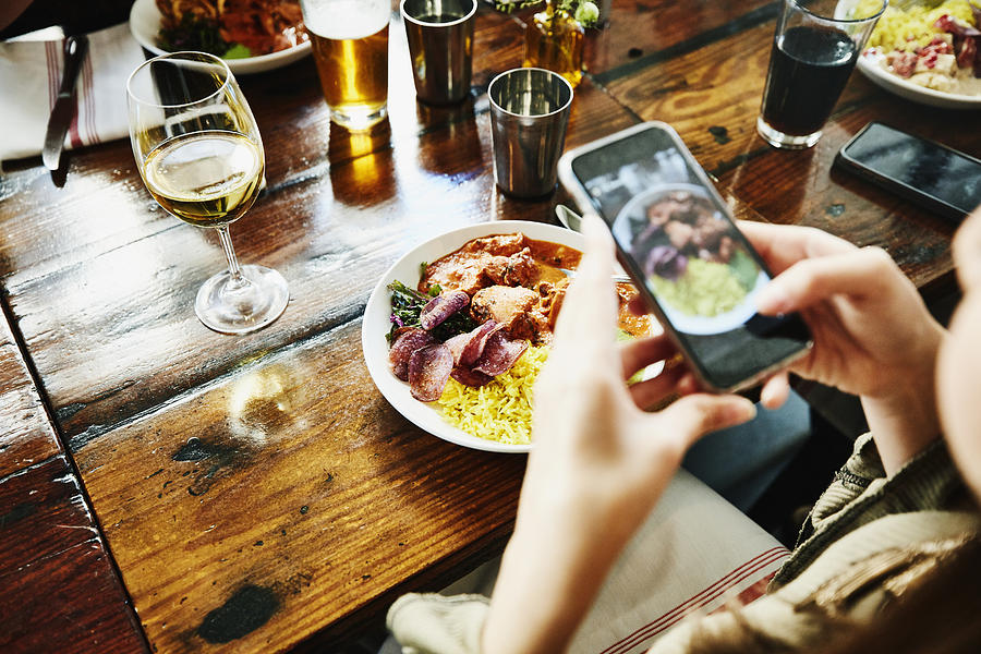 Woman taking photo of food with smartphone while having lunch with friends in restaurant Photograph by Thomas Barwick