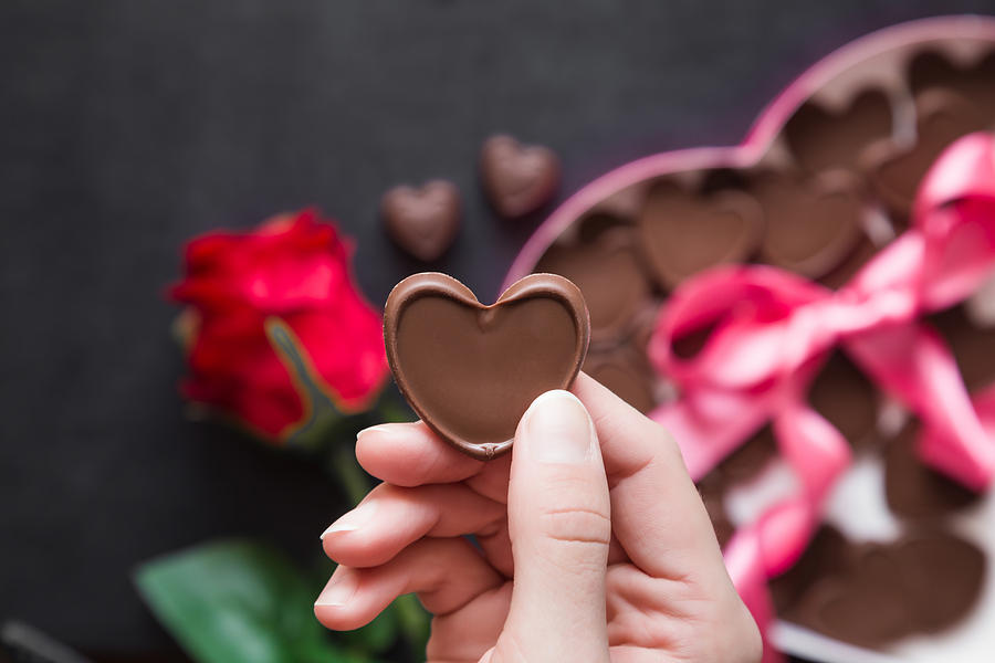 Womans hand holding a chocolate heart. Cute gift from beloved. Red rose with chocolate box of heart shape with pink ribbon on the table. Enjoying sweets. Valentines day and relationships concept. Photograph by FotoDuets