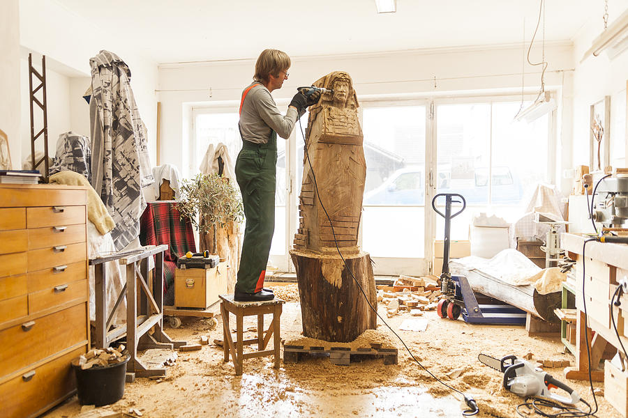 Wood carver in workshop working on sculpture with milling machine, standing on stool Photograph by Westend61