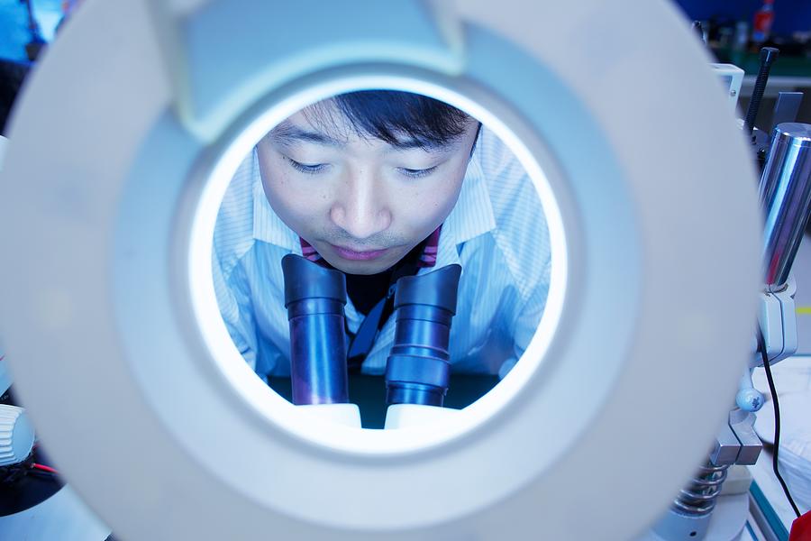 Worker at small parts manufacturing factory in China looking through microscope Photograph by Mick Ryan