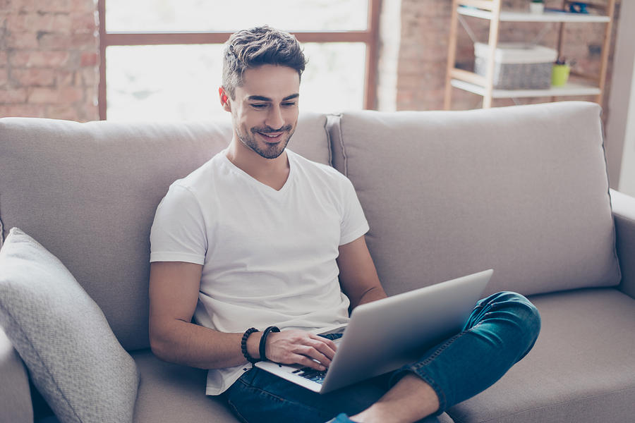Young attractive smiling guy is browsing at his laptop, sitting at home on the cozy beige sofa at home, wearing casual outfit Photograph by Deagreez