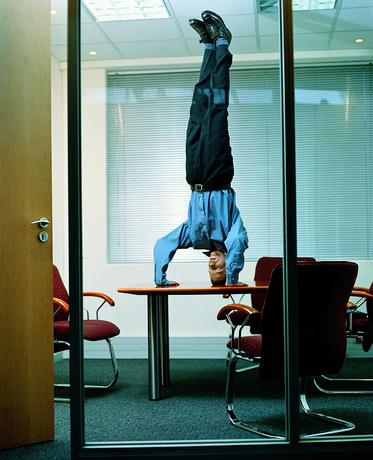 Young businessman doing headstand on office table Photograph by Daly and Newton