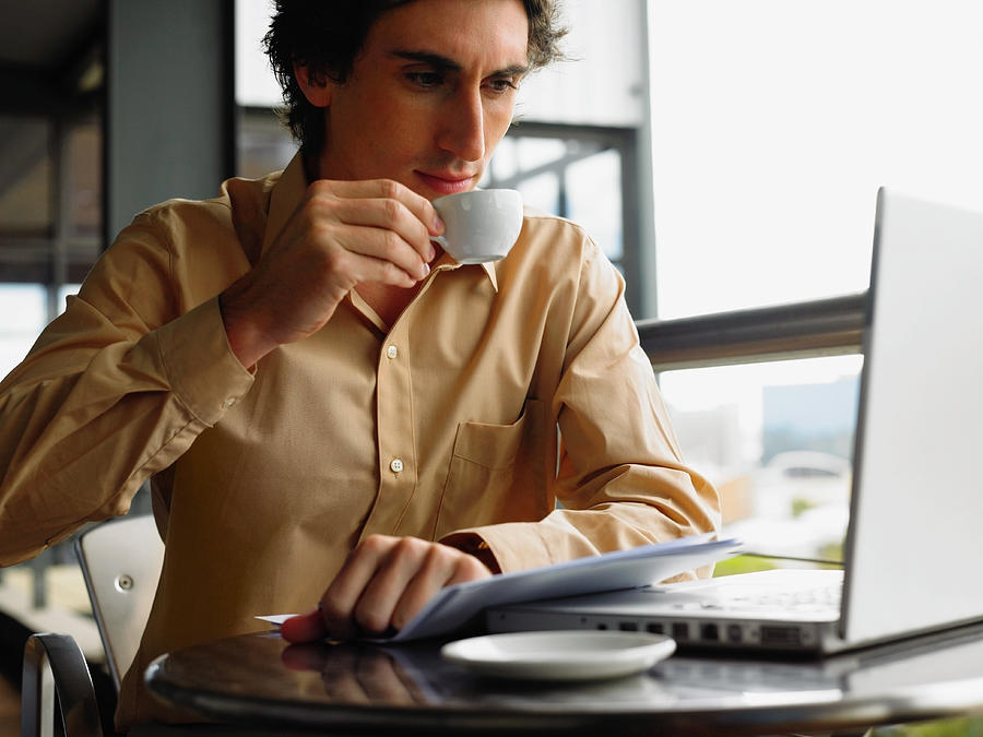 Young businessman using laptop sitting at table drinking coffee Photograph by John Rowley