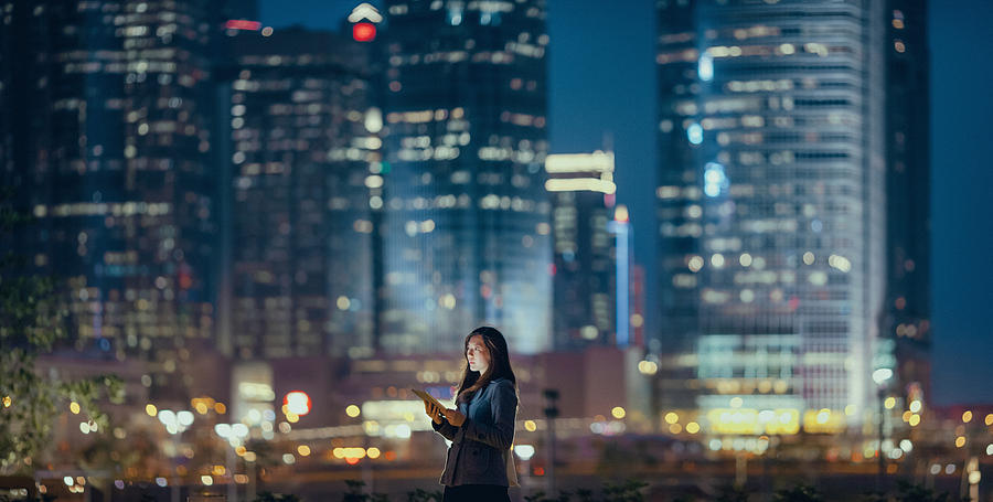 Young businesswoman using digital tablet in financial district, against illuminated corporate skyscrapers at night Photograph by D3sign