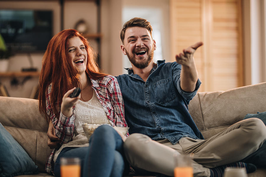 Young couple laughing while watching TV at home. Photograph by Skynesher