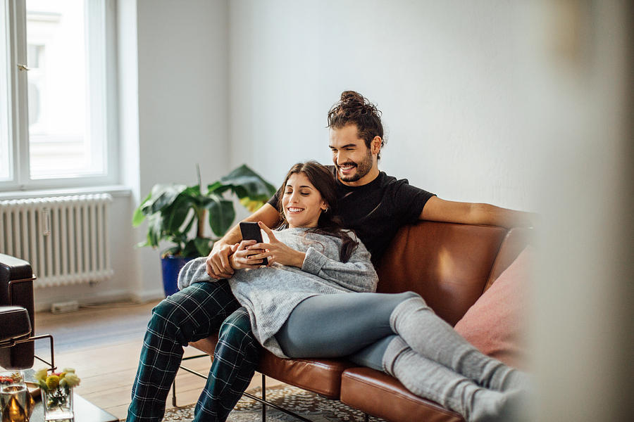 Young couple with mobile phone relaxing on sofa Photograph by Luis Alvarez