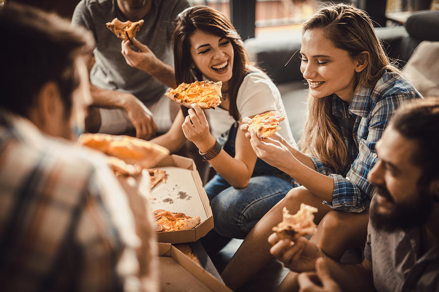 Young happy friends having fun while eating pizza at home. Photograph by Skynesher