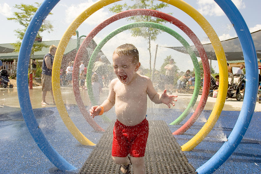 Young laughing boy runs through water park sprinklers Photograph by Kickstand