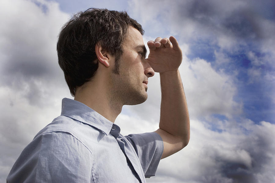 Young man against cloudy sky, shielding eyes, side view, close-up Photograph by Clandestini