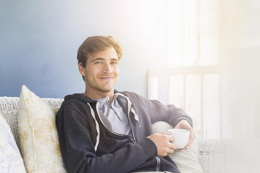Young man holding coffee cup while sitting on sofa Photograph by Portra