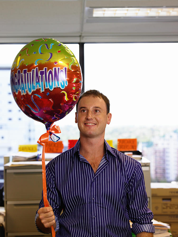 Young man holding congradulations balloon in office, smiling Photograph by David Woolley