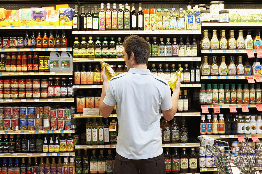 Young man in supermarket comparing bottles of oil, rear view, close-up Photograph by Noel Hendrickson