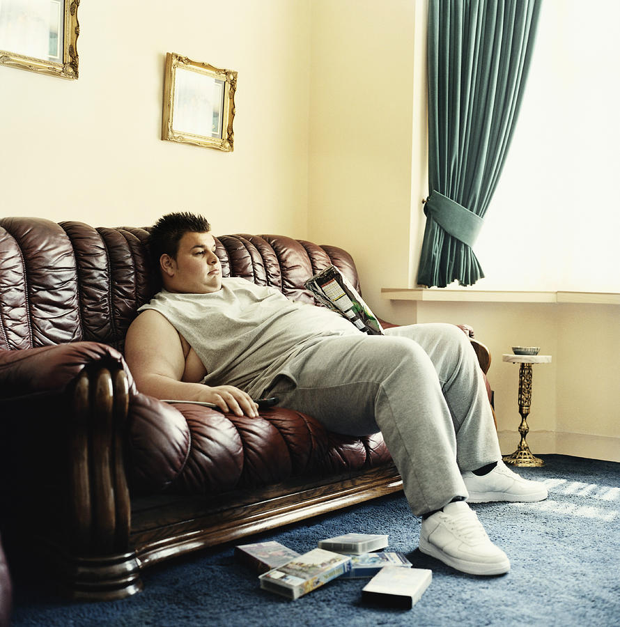 Young Man Lies Slouched on a Sofa Watching Videos and Holding a Packet of Crisps Photograph by Digital Vision.