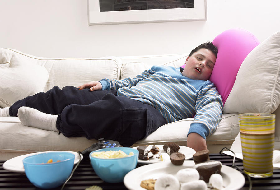 Young, Overweight Boy Sleeps on a Sofa Next to a Table of Crisps and Biscuits Photograph by Digital Vision.