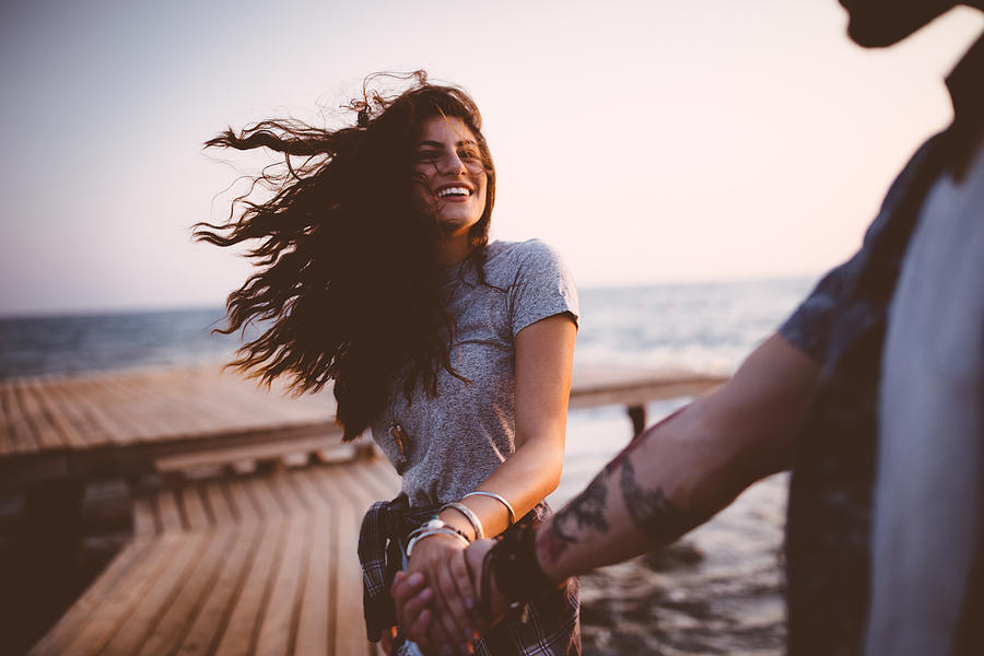 Young playful hipster couple holding hands on jetty at sunset Photograph by Wundervisuals