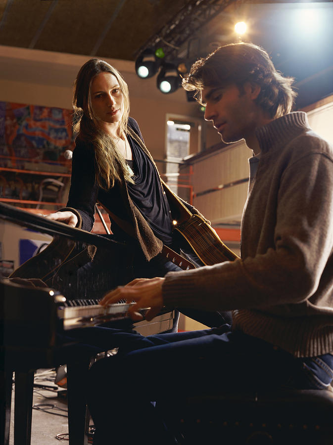 Young Woman Leaning Against a Piano as She Watches a Man Playing Photograph by Digital Vision.