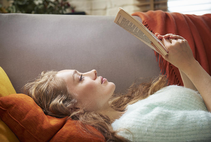 Young woman lying on sofa reading Photograph by Tara Moore