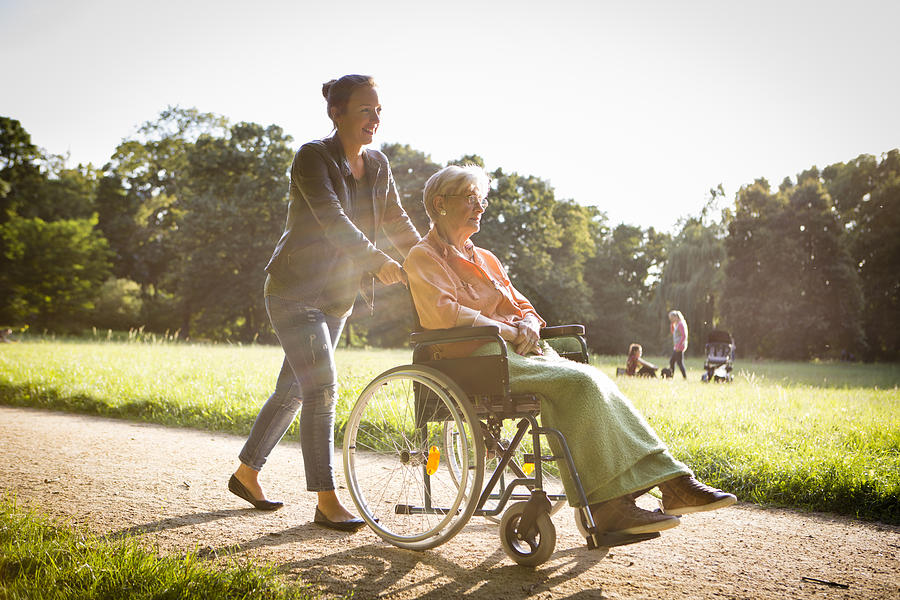 Young Woman Pushing Senior Lady In Wheelchair Through A Park Photograph by Fotografixx