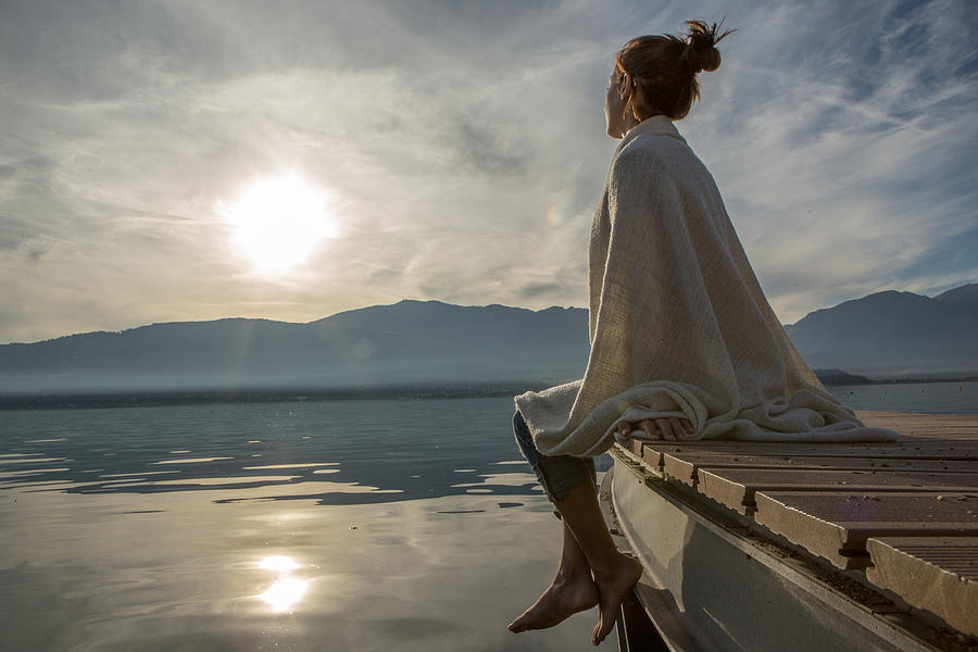 Young woman relaxes on lake pier with blanket, watches sunset Photograph by Swissmediavision