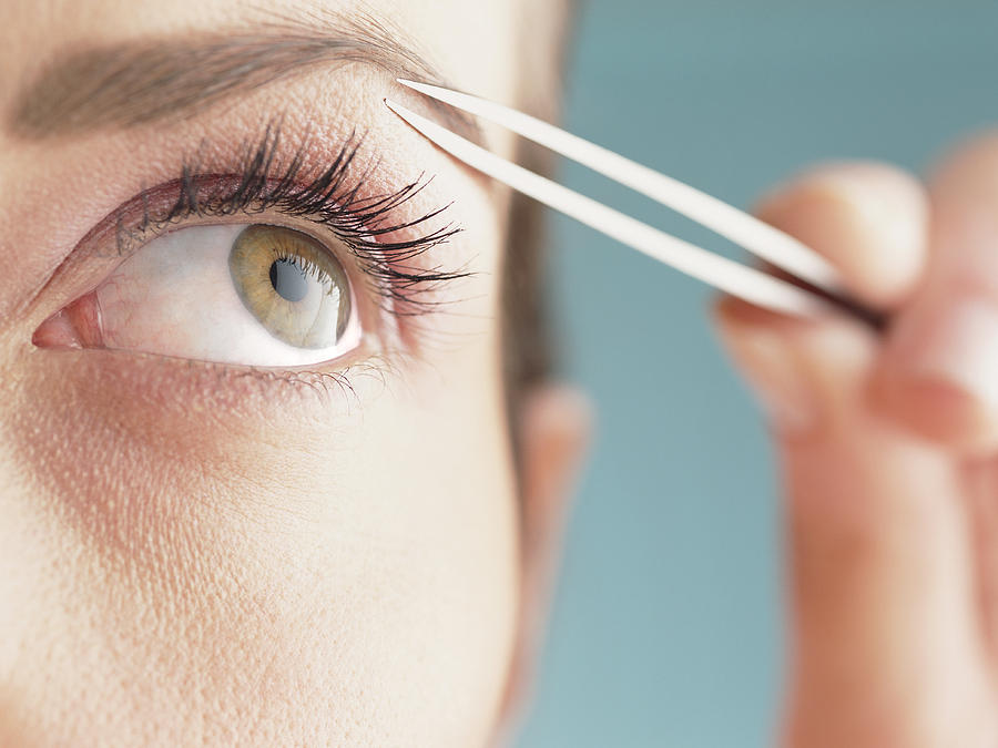 Young woman shaping eyebrow with tweezers, close-up of eye Photograph by John Slater