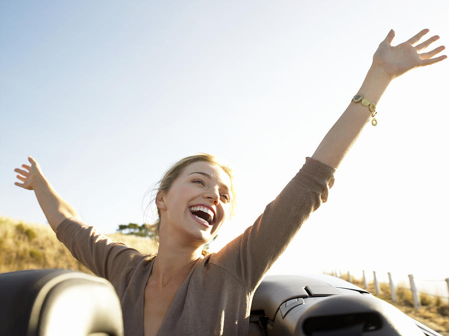 Young Woman Sits in the Back of a Convertible, Her Arms in the Air, Laughing With Joy Photograph by Digital Vision.