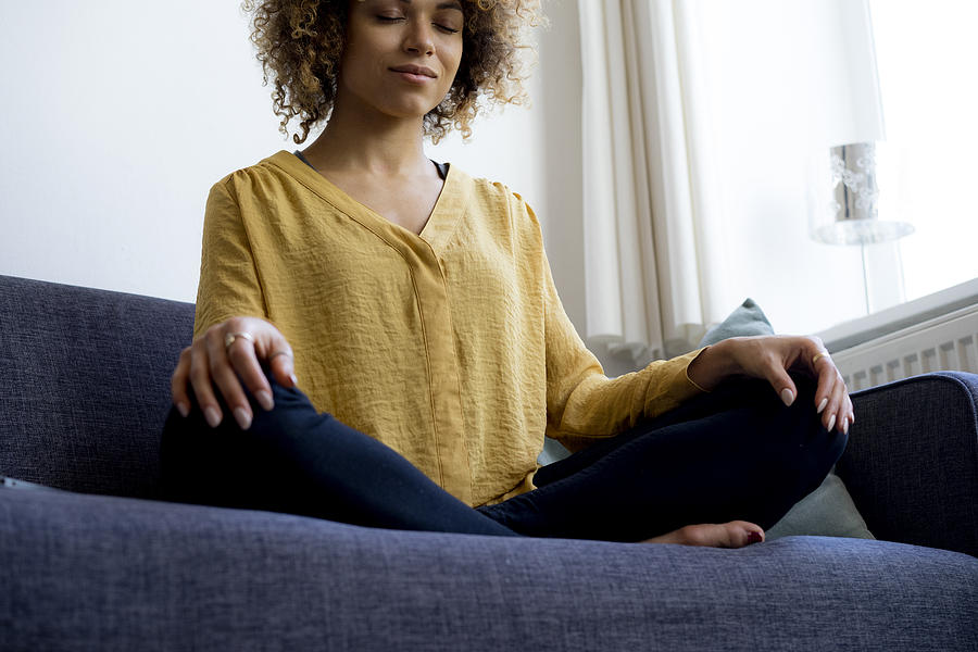Young woman sitting on couch at home meditating Photograph by Westend61