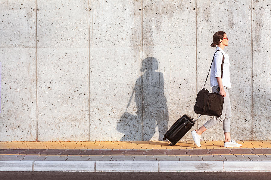 Young woman walking on a sidewalk beside the concrete wall and pulling a small wheeled luggage Photograph by Gruizza