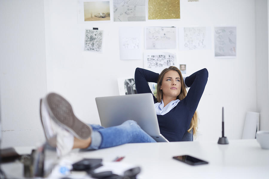 Young woman with laptop at desk in office leaning back Photograph by Westend61
