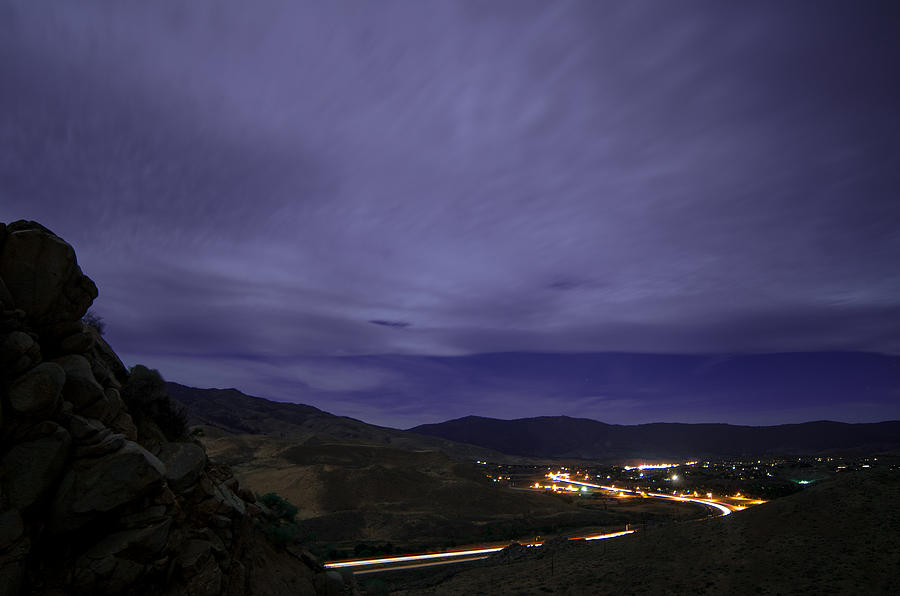 A Dramatic Purple Cloudy Sky over Dimly-Lit Mountains and Winding Vehicle Light Trails Photograph by Brian Ball