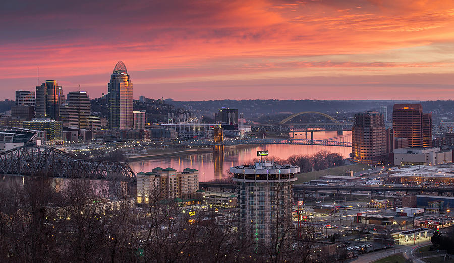 Photograph is of a view of Cincinnati from Devou Park at sunset. Many buildings have lights on that look like little twinkling stars on the ground.