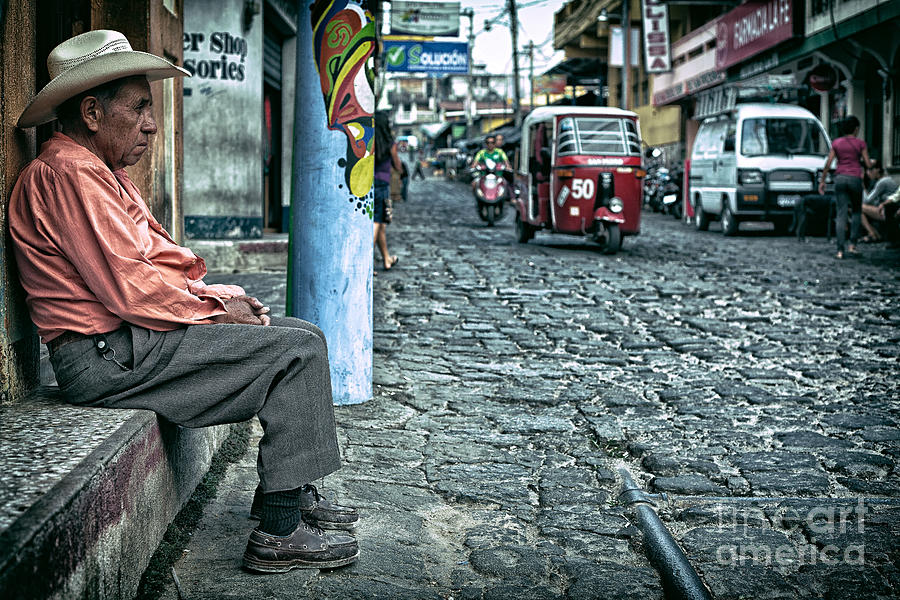 Old man sitting on an cobblestone street with traffic driving by Photograph by Sam Antonio