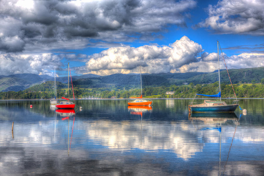Boat Photograph - Red orange and blue sailing boats on a calm lake with hills and clouds by Charlesy 