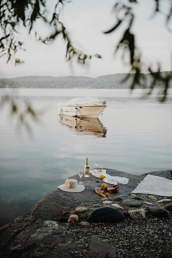 Bottle Of Sparkling Wine, Glasses, Sunhat, Sandals And Blanket On Rocky Lake Shore #1 Photograph by Agata Dimmich