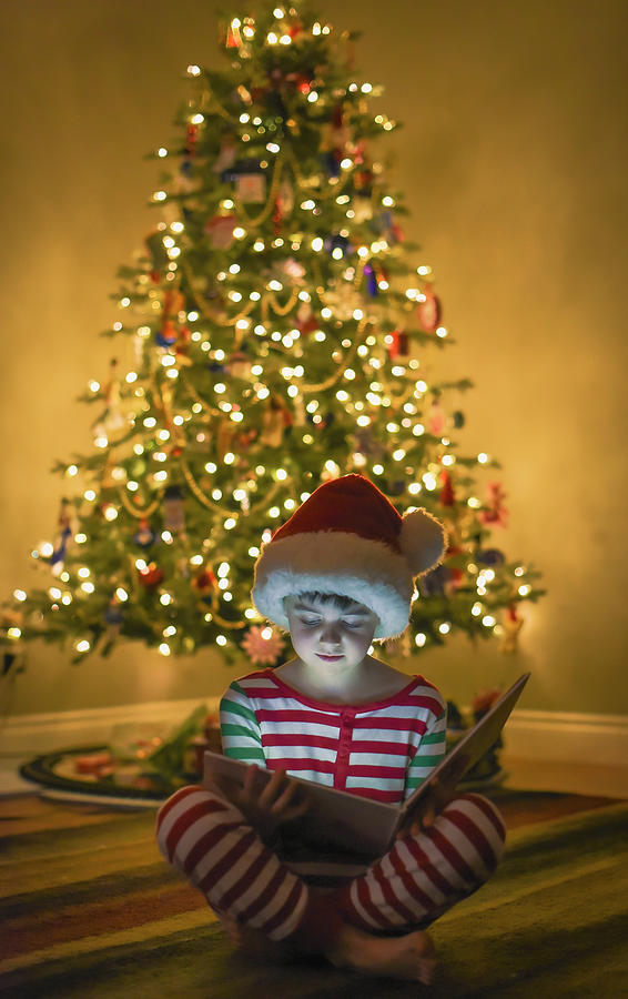Boy Reading Book In Front Of Christmas Tree With Face Illuminated