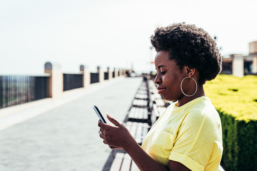 City Photograph - Portrait Of Black Girl With Afro Hair And Hoop Earrings Using Her Mobile Phone In An Urban Space In The City. #1 by Cavan Images