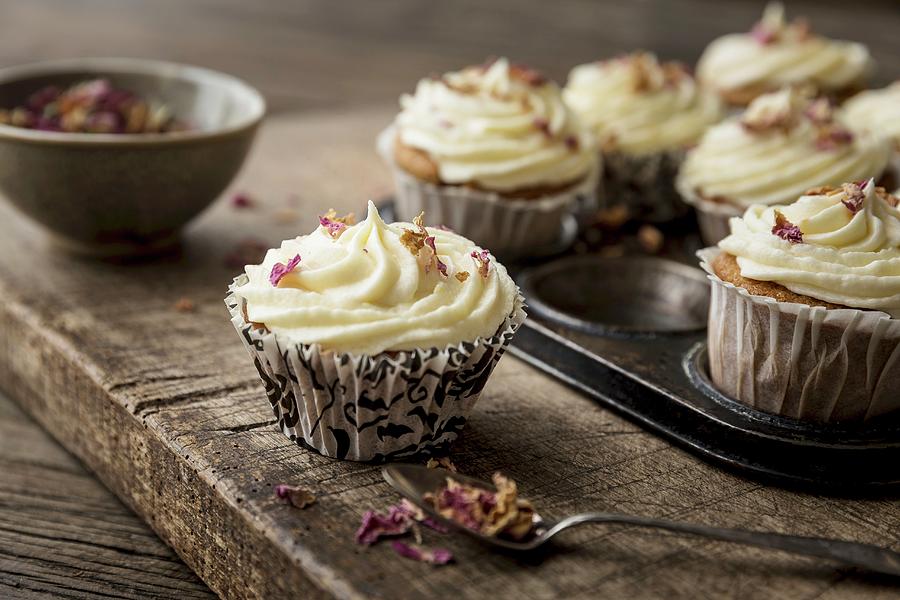 Rose And Lemon Cupcakes With Rosewater And Lemon Frosting In A Rustic Baking Tin Decorated With Dried Edible Rose Petals With Spoon And Bowl Of Rose Petals On A Wooden Board And Table #1 Photograph by Sarah Coghill