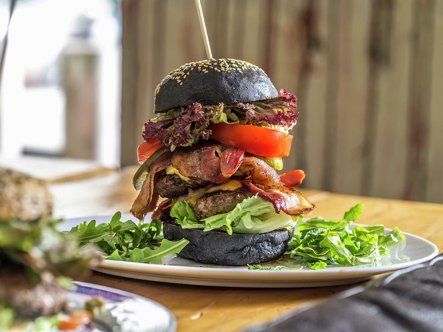 A Black Bread Double Burger With Bacon, Lettuce And Cheese On A Wooden Skewer Photograph by Manuel Krug