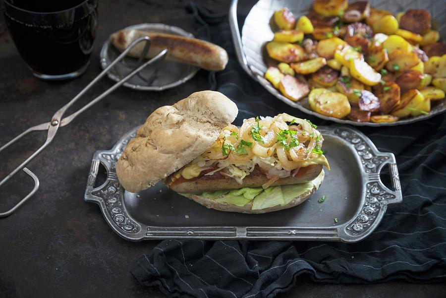 A Bread Roll With Vegan Bratwurst, White Cabbage And Roasted Onions, With A Plate Of Fried Potatoes Photograph by Kati Neudert
