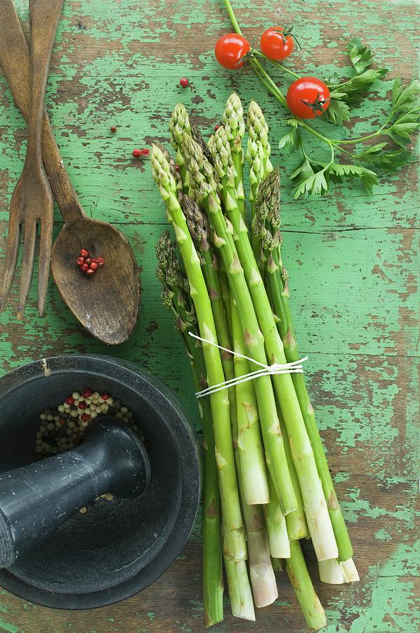 A Bundle Of Fresh Green Asparagus, Tomatoes And Parsley On A Wooden Table, Peppercorns In A Mortar And Salad Servers Photograph by Achim Sass