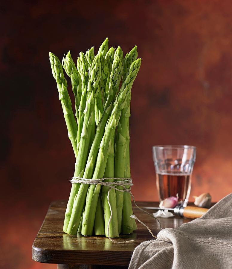 A Bundle Of Green Asparagus On A Wooden Table With Wine And Garlic Photograph by Ludger Rose