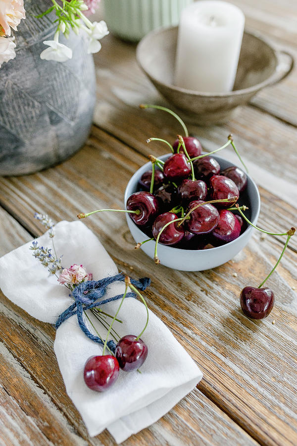 A Candle, A Vase, A Bowl Of Cherries And Fabric Napkins On A Wooden Table Photograph by Christel Harnisch