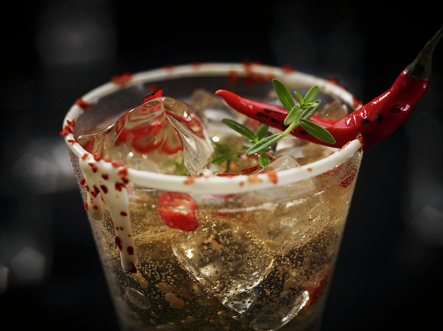 A Champagne Drink With Chilli And Ice Cubes Garnished With A Chilli Pepper And Herbs In Glass With A White Chocolate Rim Photograph by Oliver Lippert
