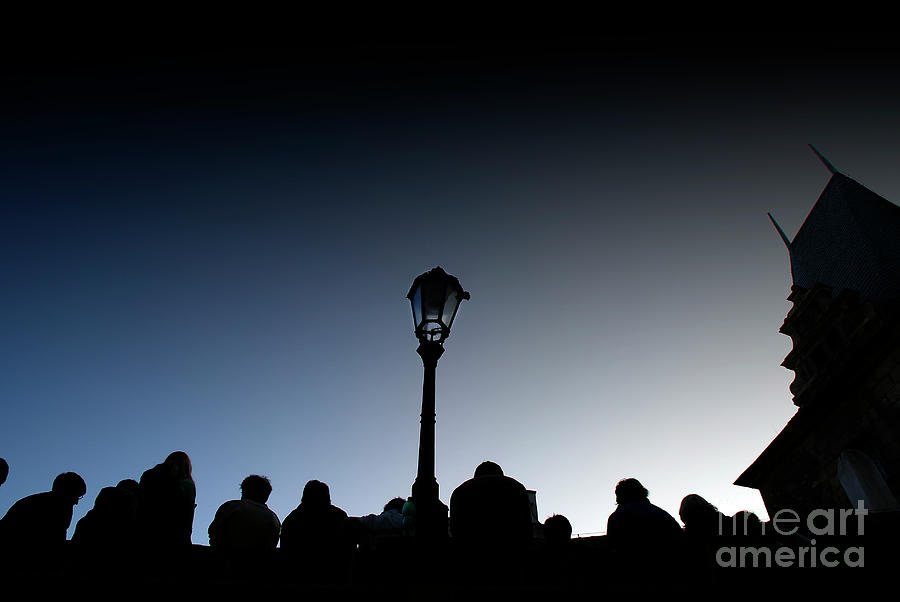  A crowd of tourists rests at dusk on a bridge, silhouetted images backlight against a streetlight. Photograph by Joaquin Corbalan