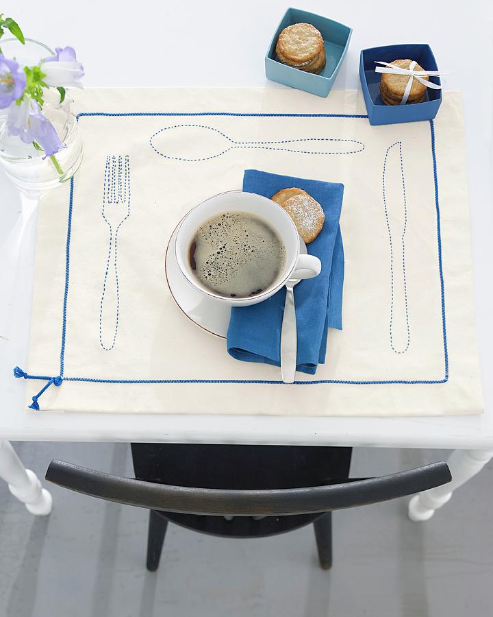 A Cup Of Coffee With Biscuits On A Decorative Place Mat With Outlines Of Cutlery And Decorative Ribbon Photograph by Jalag / Olaf Szczepaniak